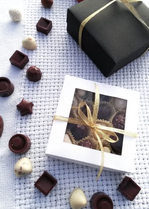 Gift box with a gold ribbon beside a windowed chocolate box on a textured light surface.