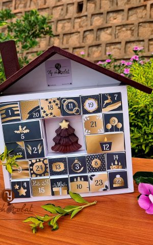 Advent calendar with numbered doors and a chocolate tree design on a wooden table, outdoors with green plants and pink flowers behind it