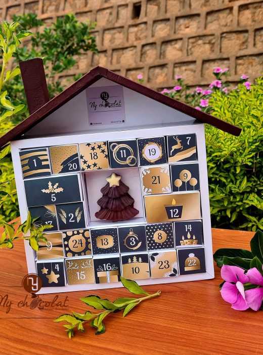 Advent calendar with numbered doors and a chocolate tree design on a wooden table, outdoors with green plants and pink flowers behind it