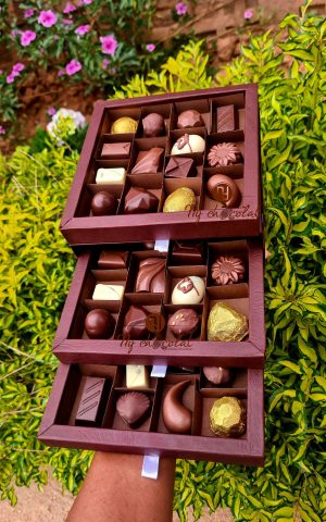 Tiered box of assorted chocolates in a maroon display outdoors among green bushes and pink flowers