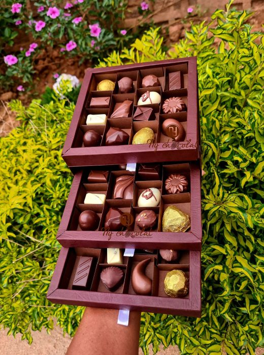 Tiered box of assorted chocolates in a maroon display outdoors among green bushes and pink flowers